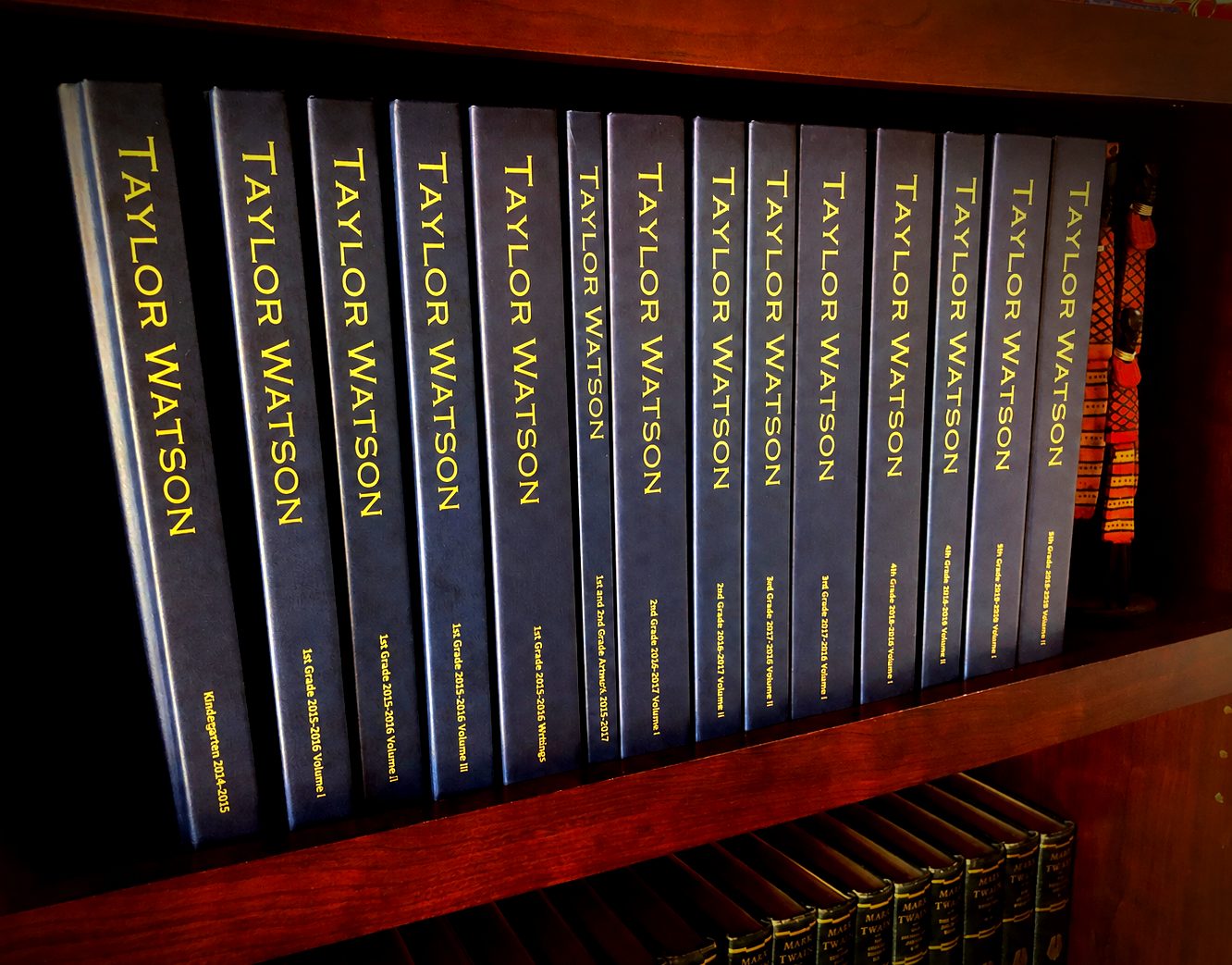 Fourteen matching navy hardcover volumes with gold foil spines — one child's set from Kindergarten through 5th grade — on a family bookshelf above a vintage Mark Twain collection